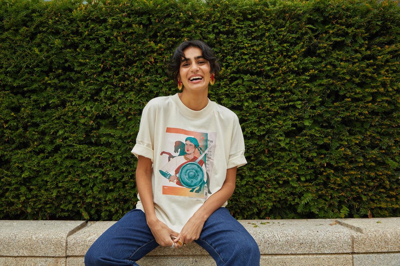 Image shows Brown person with bouncy black hair and gold earings. They sit on a stone bench with a green hedge behind them. They smile and laugh as they pose in a cream T-shirt with an graphic art image of Rani of Jhansi on the front.